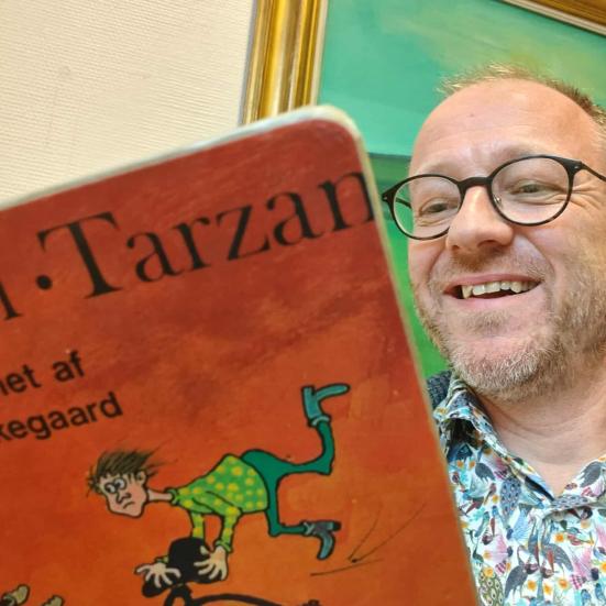 Formidler Hans Wendelboe Bøcher i spraglet skjorte læser Gummi-Tarzan. I baggrunden ses et grønt maleri.