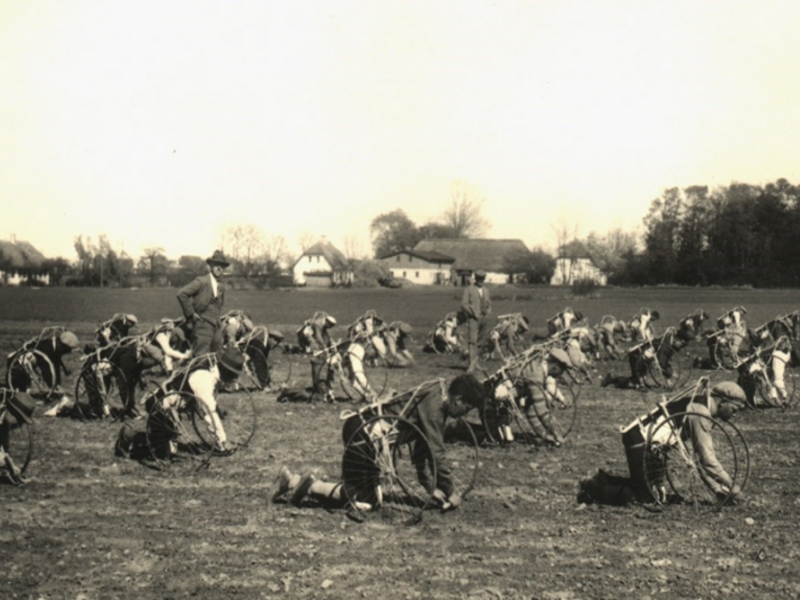 Roeentreprenør R.L.Schifters roekarle afprøver Roecyklen "STØT" på præstegårdens mark. Præstegårdsforpagter Laursen ligger midt i billedet med mørkt hår. Schifter står med hat og opfinderen Anton Olsen står med kasket i baggrunden.